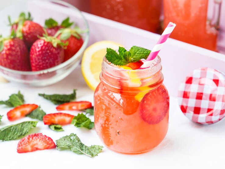 Ein alkoholfreier Erdbeer-Drink im Retro-Glas steht auf einem hellem Tablett. Drum herum liegen Erdbeere-Scheiben, eine weiße Schale mit Erdbeeren, Minzblätter sowie ein weiß-rot-karierter Deckel.