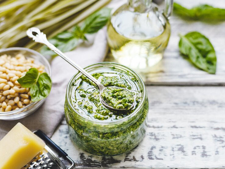 Ein Glas Mangold-Pesto mit einem Teelöffel darin, drumherum Mangoldblätter, Pinienkerne und Parmesan.