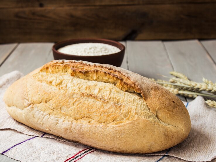 Ein selbstgebackene Brot aus vier Zutaten liegt auf einem Geschirrtuch, im Hintergrund sieht man eine Schüssel mit Mehl und etwas Getreide. | © Getty Images/ undefined undefined