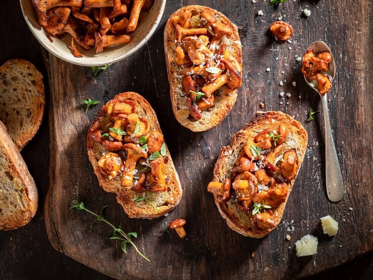 Selbstgemachte Knoblauch-Bruschetta mit Pfifferlingen liegen auf einem Untergrund aus Holz. Daneben liegen ein Löffel mit Pilzen, einige Brotscheiben und eine Schüssel mit Pilzen.