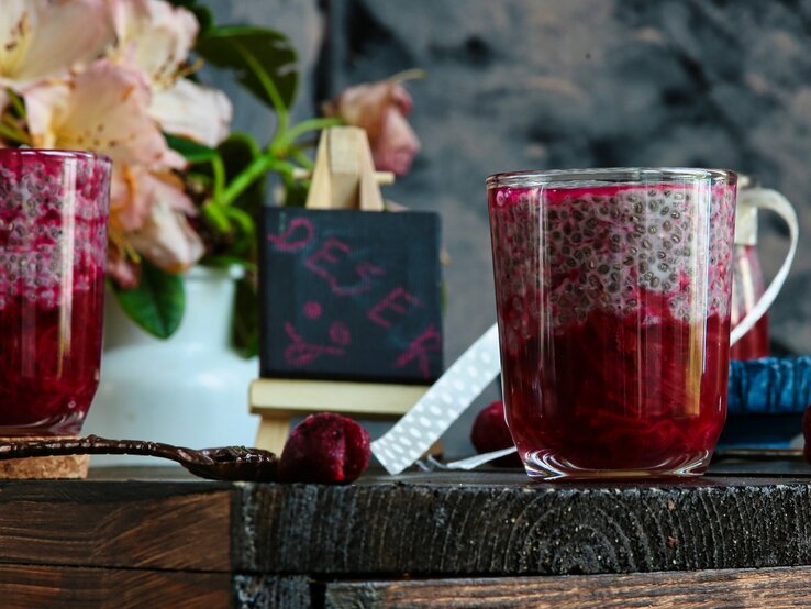 Zwei Gläser mit dem Chia-Pudding und rotem Fruchtragout auf einem Holztisch mit allerhand Deko, wie einer kleinen Tafel und ein paar Blumen.