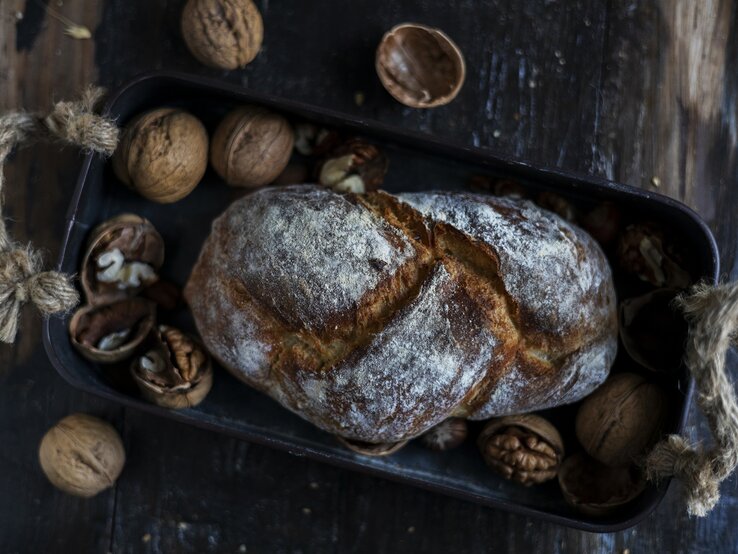 Draufsicht: Auf einem dunklen Untergrund aus Holz liegt ein Walnussbrot in einem dunklen Korb. Drumherum liegen intakte sowie geöffnete Walnüsse.