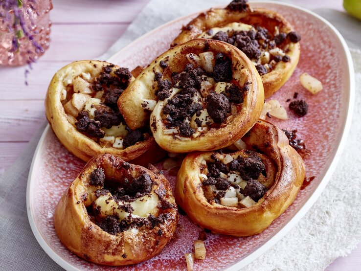 Fünf Birnen-Streuselschnecken liegen auf einer ovalen Form mit rosa Flecken. In der Form sind einige Birnenstücke und Streusel verteilt.