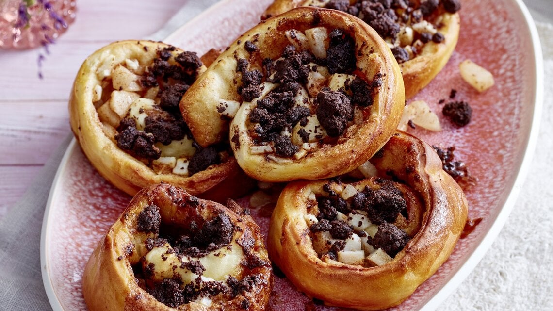 Fünf Birnen-Streuselschnecken liegen auf einer ovalen Form mit rosa Flecken. In der Form sind einige Birnenstücke und Streusel verteilt.