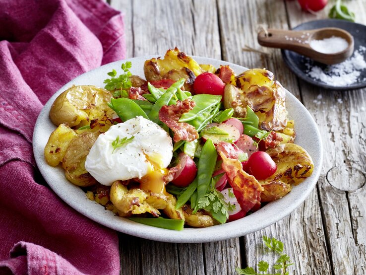 Auf einem Teller steht eine Portion Quetschkartoffeln mit Ei und Kerbel. Sie wurden mit Bacon und Radieschen verfeinert.