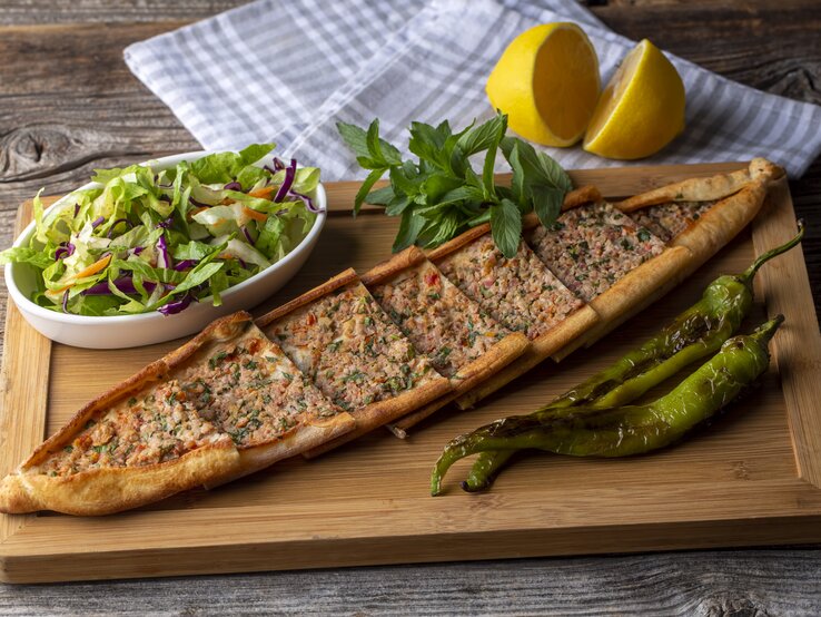 Ein Holzbrett, auf dem eine Pide mit Hackfleisch in Stücke geschnitten wurde. Daneben steht eine Schale Salat, außerdem liegen daneben eine aufgeschnittene Zitrone, ein Geschirrtuch, grüne Chilis und Kräuter.