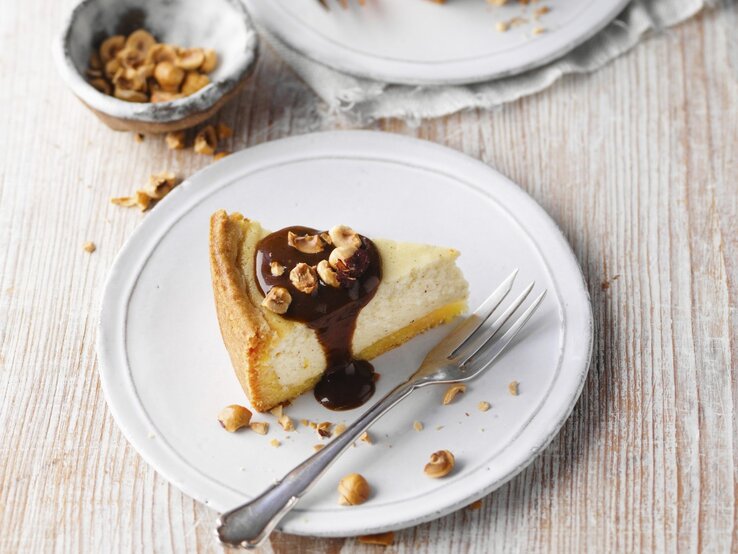 Unser rheinischer Käsekuchen, auch bekannt als Klatschkiestaat, ist genau der Richtige für Käsekuchen-Fans. Auf einem Teller ist ein Stück Klatschkiestaat, ein rheinischer Käsekuchen, serviert. Er ist mit Zuckerrübensirup und Haselnüssen getoppt.