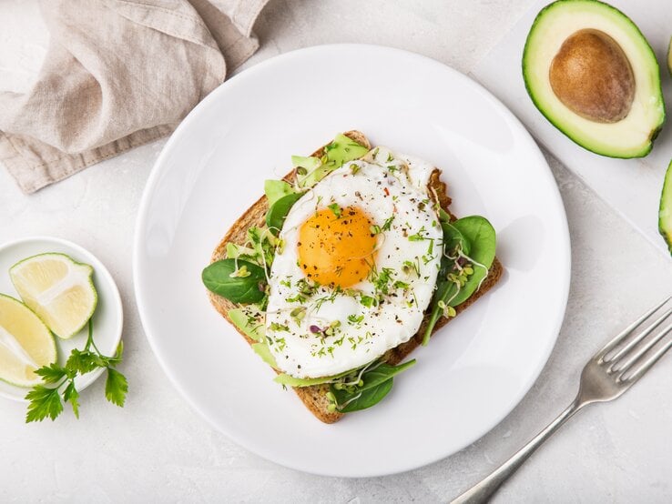 Ein Toast mit Avocado, Salat und Spiegelei liegt auf einem weißen Teller auf einem hellen Tisch. Rechts neben dem Teller liegt eine halbierte Avocado und eine Gabel. Links neben dem Teller steht eine kleine Schale mit Limettenstücken. Am oberen Bildrand liegt ein beiges Tuch.