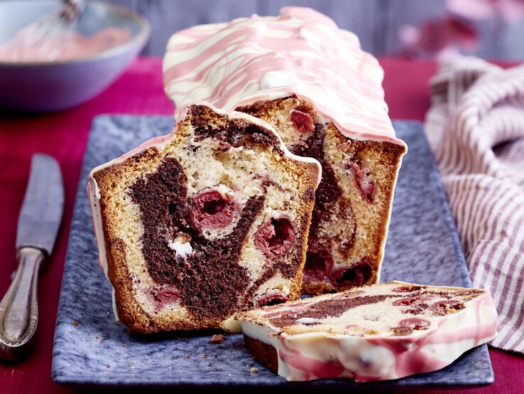 Auf einem langen Teller steht ein angeschnittener Marmor-Kirschkuchen aus der Kastenform. Er ist verziert mit einer Glasur aus weißer Schokoladen mit rosa Linien. 