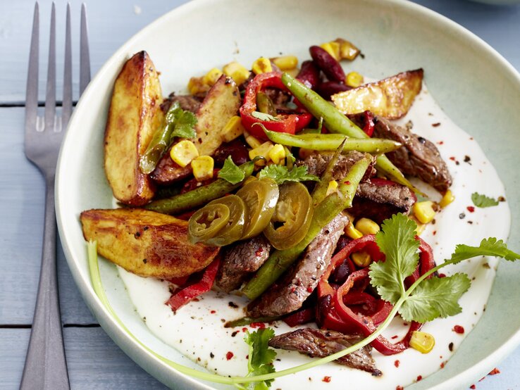 Mexikanisches Geschnetzeltes mit angerösteten Bohnen, Kartoffeln, Mais, Chili und frischem Koriander, serviert auf einem weißen Teller auf hellblauem Holztisch. 