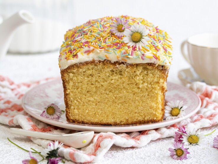 Auf einem Teller liegt ein Vanillekuchen mit Zuckerglasur und bunten Zuckerstreuseln aus der Kastenform. Er ist dekoriert mit essbaren Blüten und Zuckerstreuseln. 