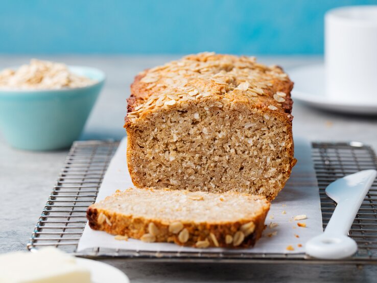 Bananenbrot mit Haferflocken auf Kuchenrost, dahinter eine blaue Schüssel mit Haferflocken.