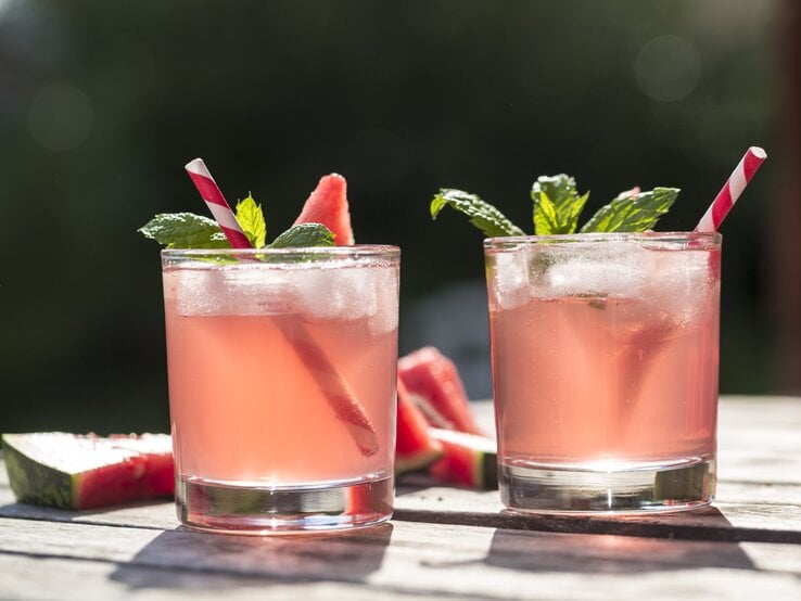 Zwei Gläser gefüllt mit rosafarbenem Wasser-Melonen-Hugo – Dekoriert mit Frucht, Minze und Strohalmen. 