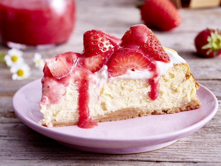 Auf einem Teller ist ein Stück Vanille-Erdbeer-Cheesecake serviert. Der Hintergrund ist hölzern und Gänseblümchen sind darauf verteilt. 