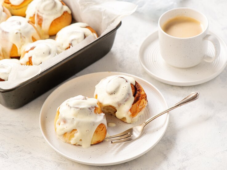 Zimtbrötchen mit Glasur in Auflaufform und auf Teller neben einer Tasse Kaffee. Alles angerichtet auf einer weißen Tischdecke.