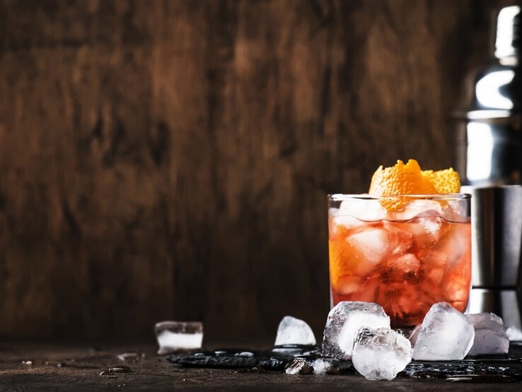 Ein Glas Aperol Sour und ein Cocktail-Shaker stehen in einem Raum aus Holz. Daneben liegen Eiswürfel. 