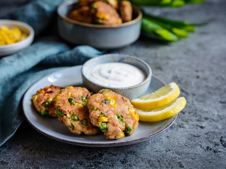 Mehrere Thunfisch-Frikadellen mit Erbsen, Mais und Frühlingszwiebeln auf einem Teller, serviert mit einem frischen Sauerrahmdip und Zitronenscheiben. 