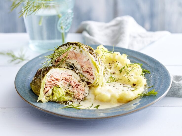 Lachs-Wirsing-Wickel, garniert mit Dill und serviert mit Kartoffelpürree auf einem Tonteller auf weiß gedecktem Tisch. 