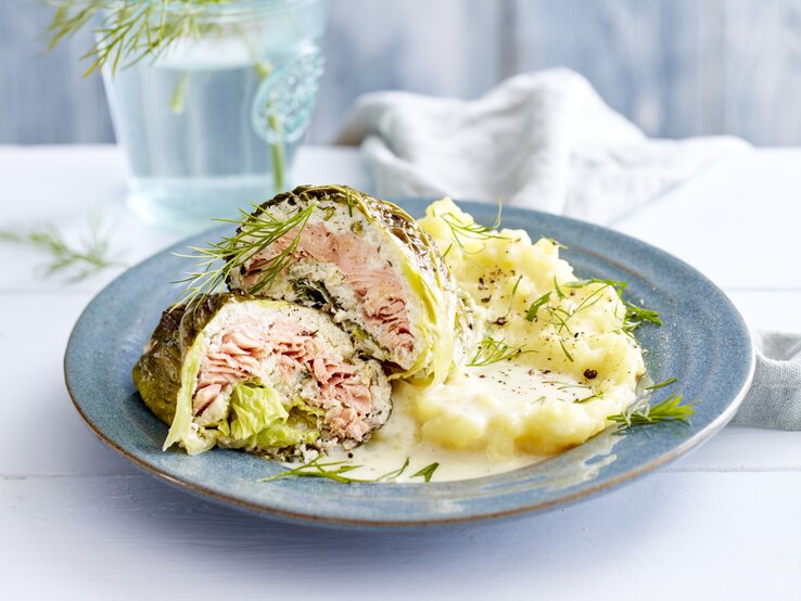 Lachs-Wirsing-Wickel, garniert mit Dill und serviert mit Kartoffelpürree auf einem Tonteller auf weiß gedecktem Tisch. 