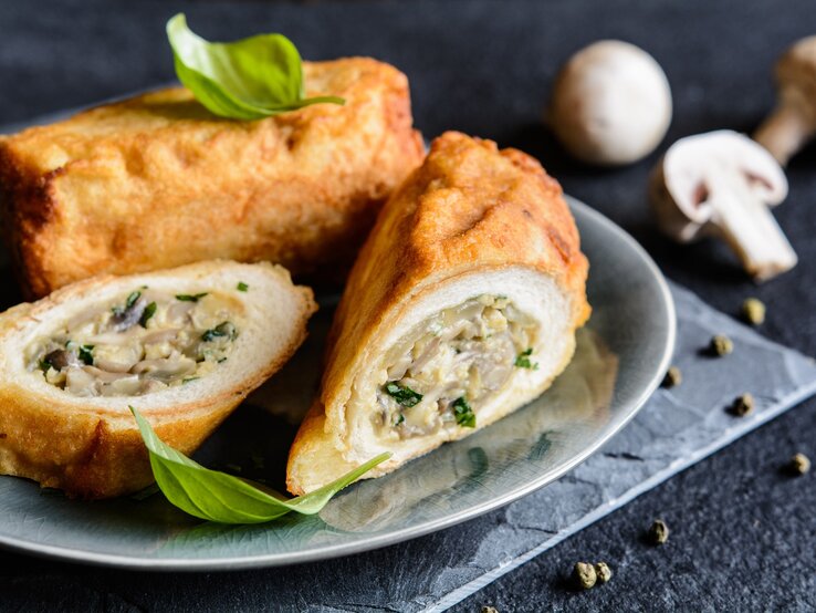 Toastrollen mit Pilzen und Käse auf grauem Teller, grauem Tuch und dunklem Tisch. Eine Rolle im Ganzen, eine aufgeschnitten dem Betrachter zugewandt. Basilikumblätter als Deko oben auf den Rollen. Im Hintergrund weiße Champignons. Frontalaufnahme.