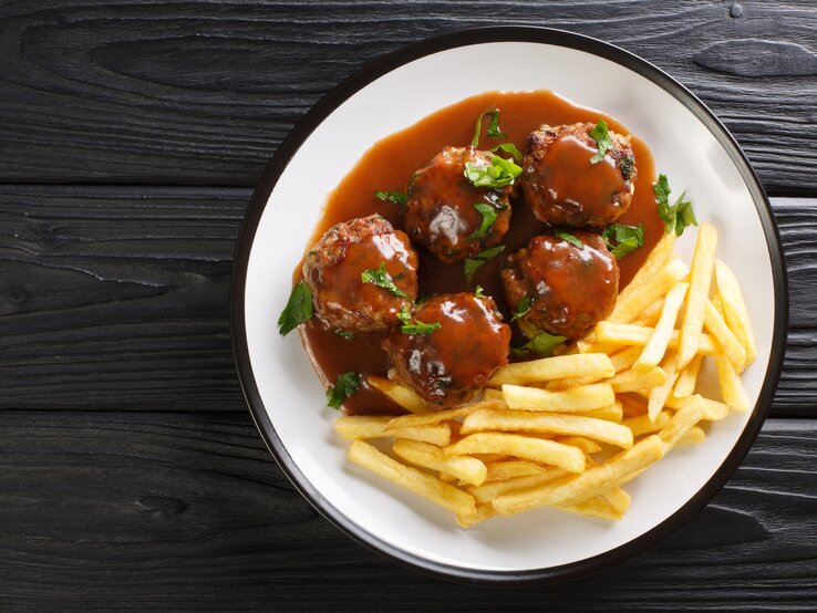 Lütticher Buletten sind ein Klassiker in Belgien. Muss jeder mal gegessen haben! Lütticher Buletten und Pommes auf weißem Teller, angerichtet auf dunklem Holz.