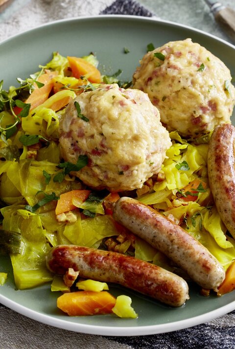 Auf einem Teller ist eine Portion Speckknödel auf geschmortem Weißkohl angerichtet. Dazu gibt es Rostbratwürstchen. 