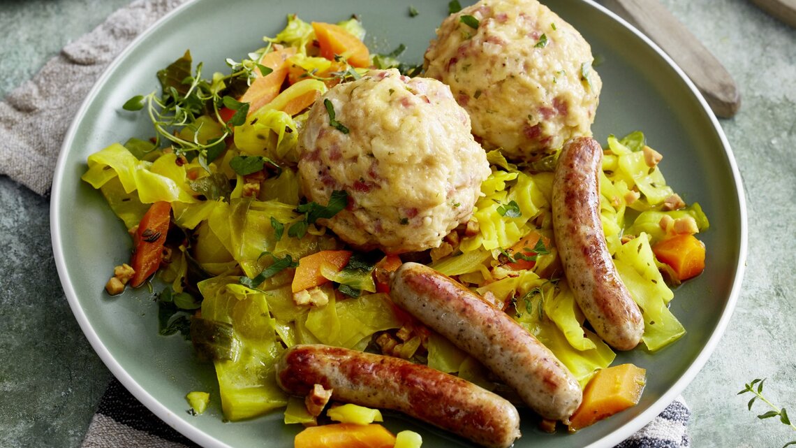 Auf einem Teller ist eine Portion Speckknödel auf geschmortem Weißkohl angerichtet. Dazu gibt es Rostbratwürstchen. 