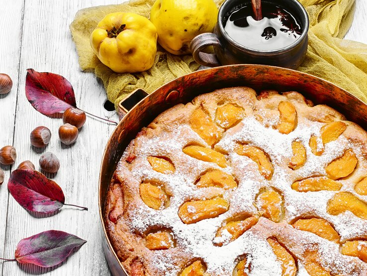 Perfektes Landfrauen-Rezept: Ein französischer Quittenkuchen! Draufsicht: ein runder Kuchen mit Quitten, der noch in der Backform ist. Er liegt auf einem hellen Untergrund aus Holz. Daneben liegen Blätter von einem Baum, Haselnüsse und Quitten. Neben dem Kuchen steht eine dunkle tasse mit einer dunklen Flüssigkeit.