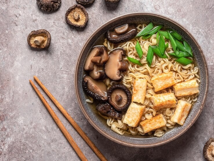 Eine Schüssel schnelle Ramen mit Nudeln, Tofu, Pilzen und Frühlingszwiebeln in der Draufsicht, darum herum liegen Pilze und Essstäbchen.