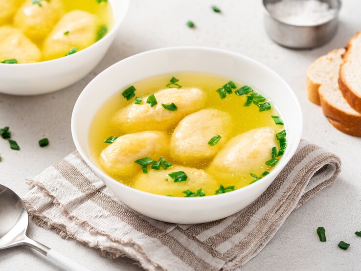 In Bayern heilig, in ganz Deutschland beliebt: Eine bayerische Grießnockerlsuppe. Draufsicht, schräg: Zwei weiße Schüsseln mit einer bayerischen Grießnockerlsuppe stehen auf einem hellen Untergrund. Daneben liegen ein Löffel, zwei Scheiben Weißbrot und eine Schale mit Salz.
