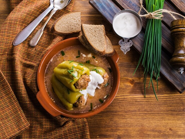 Gefüllte Paprika ungarischer Art mit Soße in einer Tonschüssel auf einem Holztisch mit Brot und frischem Schnittlauch.