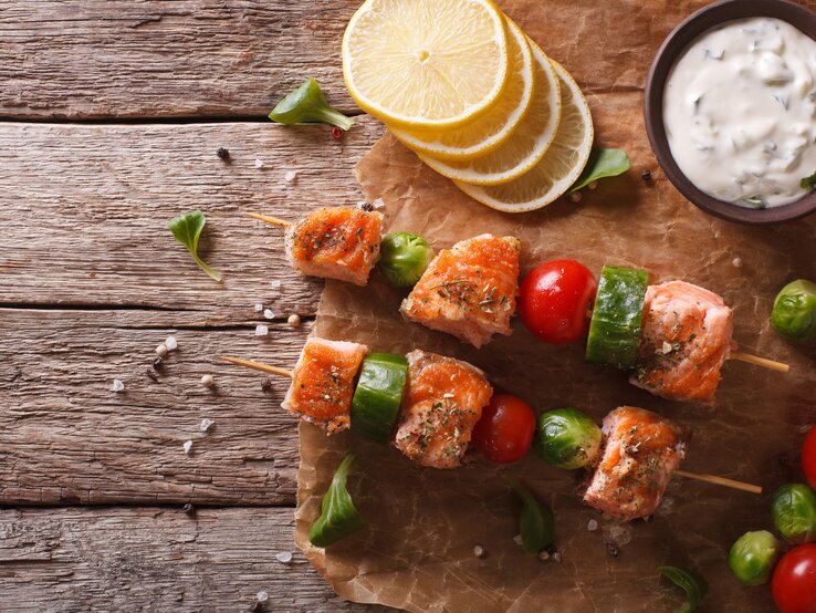 Draufsicht: Zwei Rosenkohl-Lachs-Spieße liegen auf Papier und einem Untergrund aus Holz. Daneben liegen Zitronenscheiben, Rosekohl-Röschen und eine Schale mit Kräuterquark.