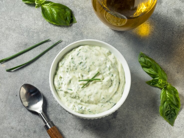 Eine Schale mit Kräuter-Mayonnaise. Daneben steht eine Karaffe Öl und einige Kräuter liegen daneben.