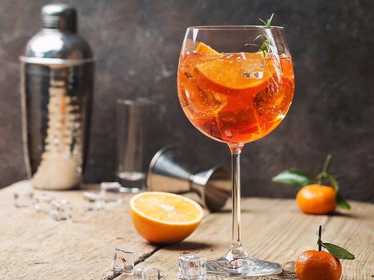 Ein Glas alkoholfreier Aperol Spritz mit Orangenscheiben und Eis, dahinter Cocktailzubehör.