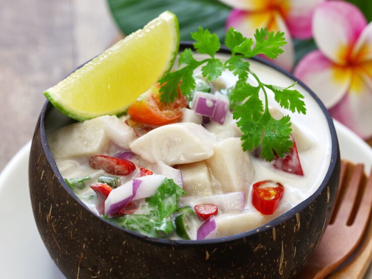 Wunderschön und himmlisch sommerlich. Unsere Ceviche mit Kokosmilch, Chili und frischem Gemüse ist ein absoluter Traum. Ceviche mit Kokosmilch und Chili in Kokosschale auf hellem Untergrund. Daneben Blütenarrangement. Draufsicht.