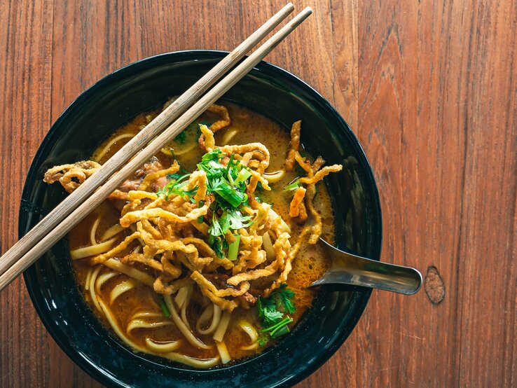 Eine dunkle Schale Khao Soi auf einem dunklen Holztisch mit zwei Holzstäbchen.