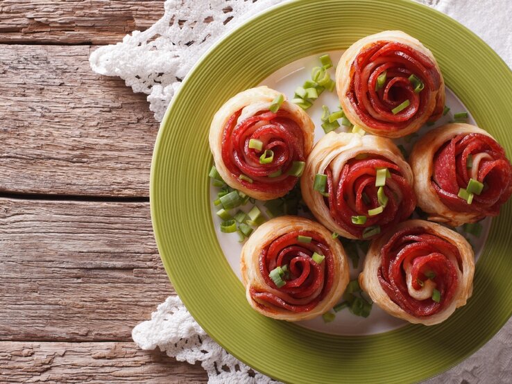 Mehre Salami-Blätterteig-Rosen auf einem weißen Teller mit grünem Rand und rustikalem Holztisch. Darunter noch eine weiße Tischdecke und alles direkt von oben fotografiert.