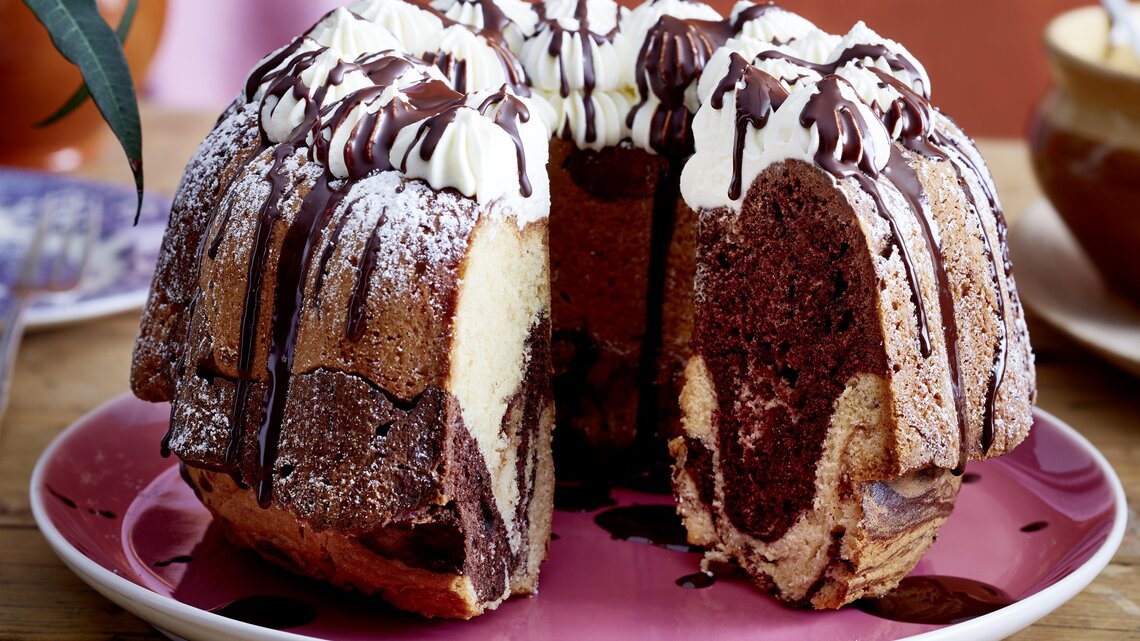 Auf einem Teller liegt ein angeschnittener Baileys-Marmorkuchen. Er ist mit Sahnetüpfeln verziert und mit Schokolade überzogen. 