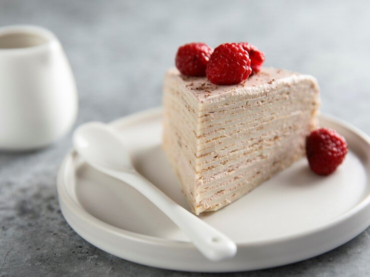 Auf einem Teller liegt ein Stück des Himbeer-Crepe-Kuchen. Man erkennt die Pfannkuchen-Schichten und die Cremefüllung. Er ist dekoriert mit Himbeeren.