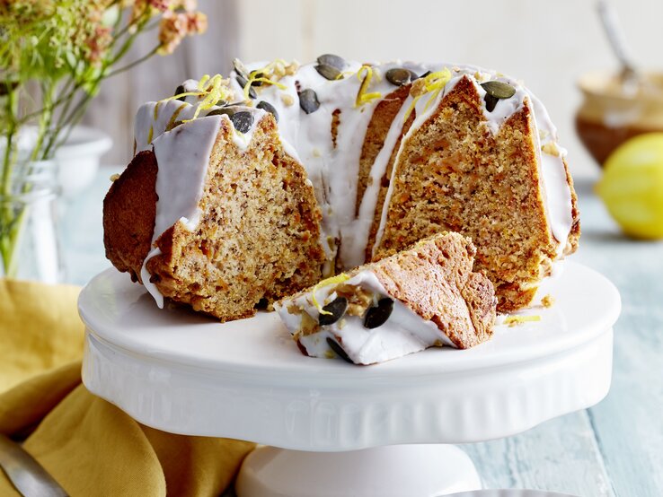 Auf einem Porzellan-Teller steht ein angeschnittener Möhren-Walnuss-Gugelhupf. Er ist mit Zuckerguss überzogen und mit Kürbiskernen verziert.