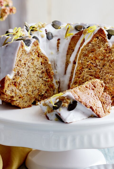 Auf einem Porzellan-Teller steht ein angeschnittener Möhren-Walnuss-Gugelhupf. Er ist mit Zuckerguss überzogen und mit Kürbiskernen verziert.