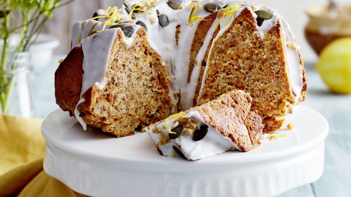 Auf einem Porzellan-Teller steht ein angeschnittener Möhren-Walnuss-Gugelhupf. Er ist mit Zuckerguss überzogen und mit Kürbiskernen verziert.