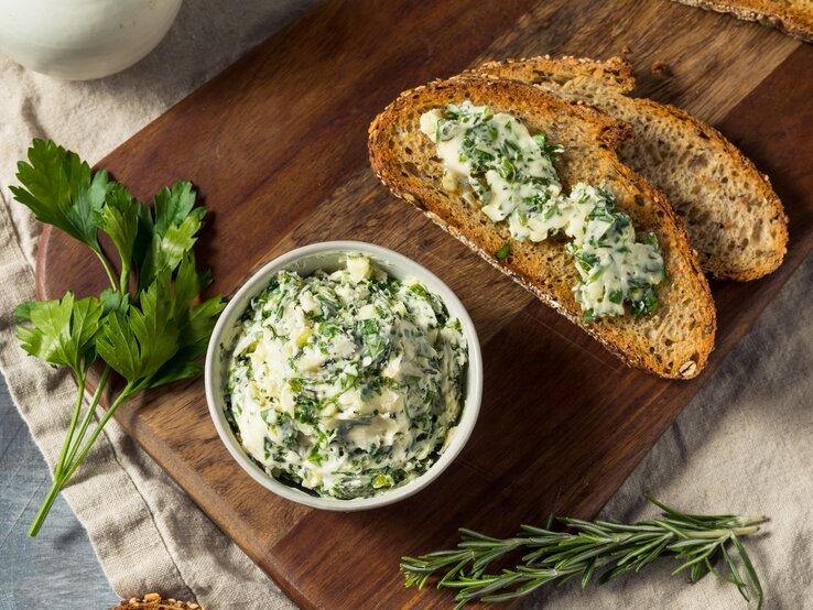 Eine Schale vegane Kräuterbutter, daneben zwei Brotscheiben mit der Butter bestrichen, außerdem frische Kräuter. Draufsicht.