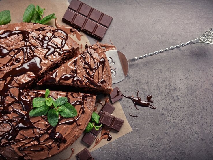 Schokoladentorte mit Minzblättern auf modernem, grauem Untergrund mit Tortenheber und Schokoladentafeln. 