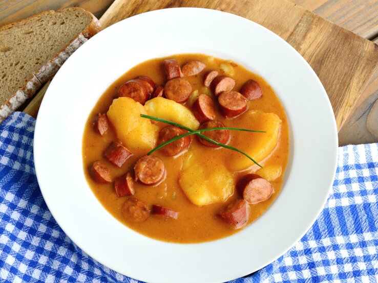 Ein weißer Teller mit der Suppe auf einem Holztisch mit einer blau-weiß-karierten Decke. Alles von schräg oben fotografiert.