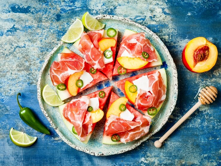 Eine Scheibe Wassermelone unterteilt in sechs gleichgroße Pizzastücke belegt mit Feta, Prosciutto, Jalapeños und Aprikosen, neben Limetten Stücken, von oben, vor einem blauen Hintergrund. 