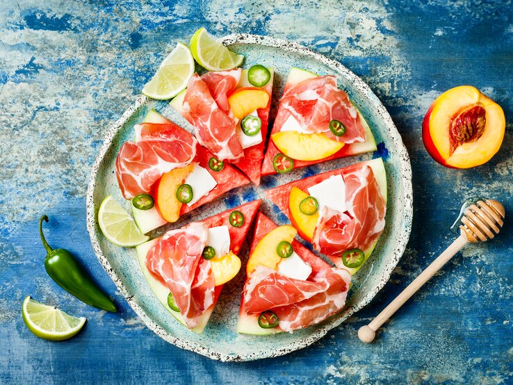 Wassermelonen-Pizza-Feta-Prosciutto Eine Scheibe Wassermelone unterteilt in sechs gleichgroße Pizzastücke belegt mit Feta, Prosciutto, Jalapeños und Aprikosen, neben Limetten Stücken, von oben, vor einem blauen Hintergrund.