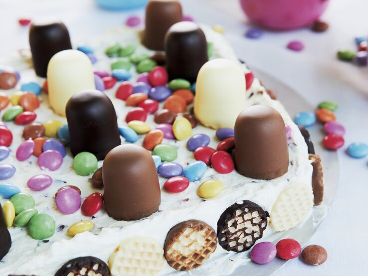 Auf einem Teller liegt eine bunte Mini-Schokokuss-Torte. Sie ist dekoriert mit Schokoküssen und Smarties. Im Hintergrund sind zwei Tassen zu sehen. 
