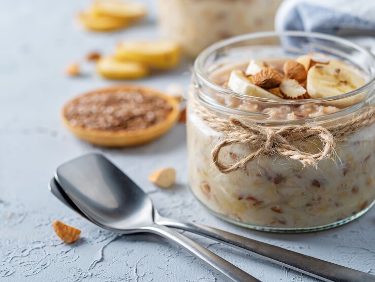Ein Glas mit dem Leinsamen-Pudding auf hellblauem Untergrund. Daneben noch unscharf gehackte Nüsse und Bananenscheiben.