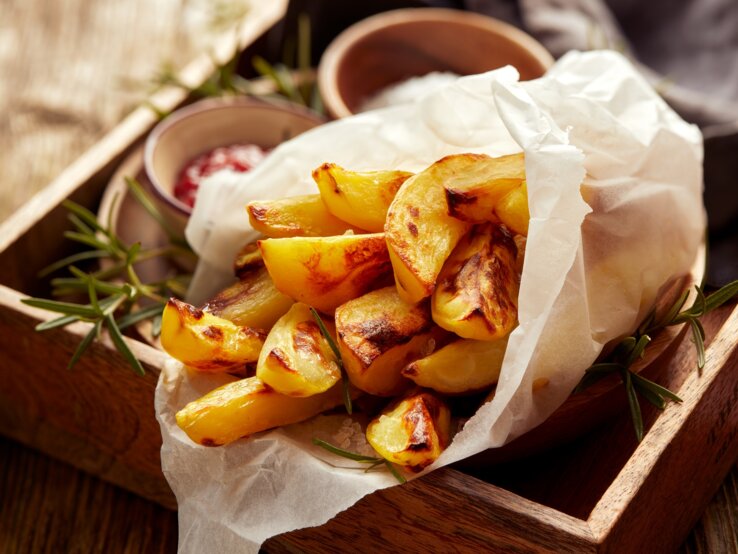 Eine Portion Kohlrabi-Pommes, eingewickelt in eine Papiertüte, auf einem Holztablett. Daneben Schälchen mit Dips.